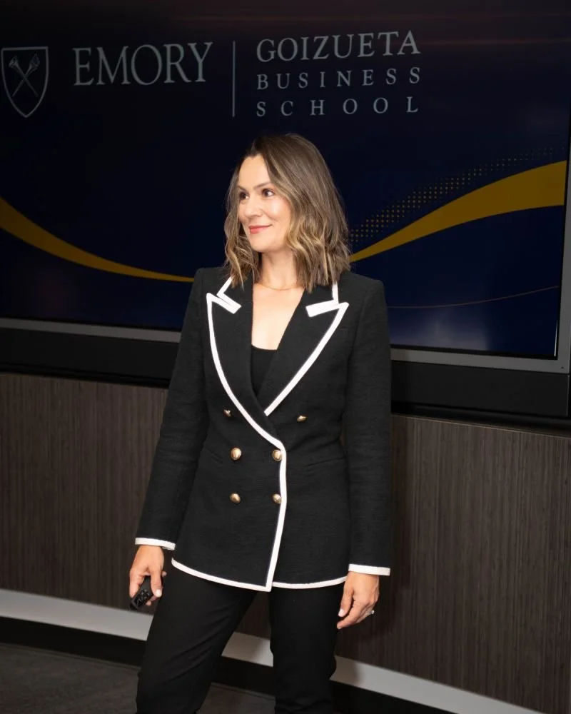 Marina Cooley in a black blazer with white trim and gold buttons standing in front of a screen displaying 'Emory Goizueta Business School' logo.