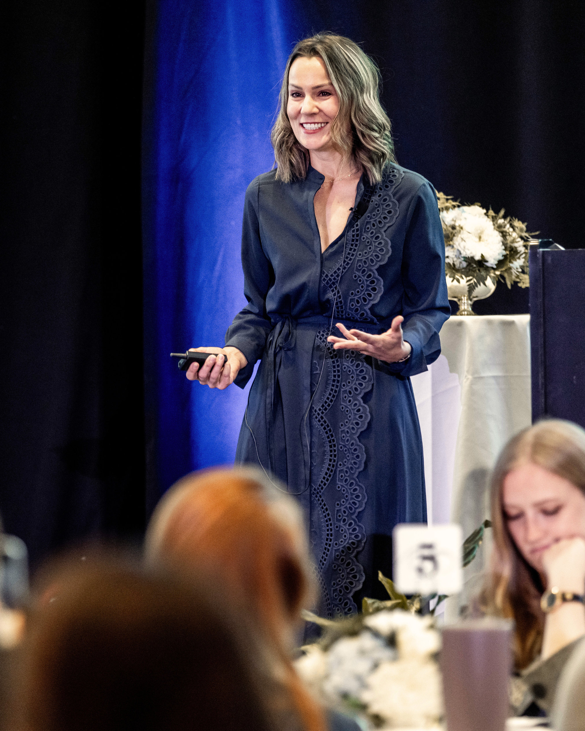 Marina Cooley pictured smiling while speaking at an event with a dark background and blue lighting, with a table of flowers and people in the foreground.