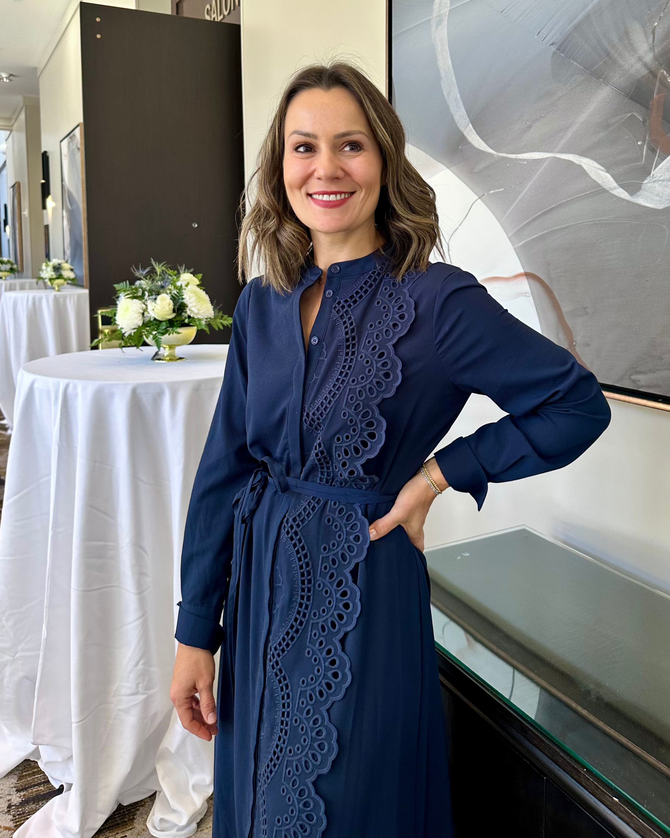 Marina Cooley wearing a navy blue dress with lace details, smiling and posing indoors near a table with a floral arrangement.