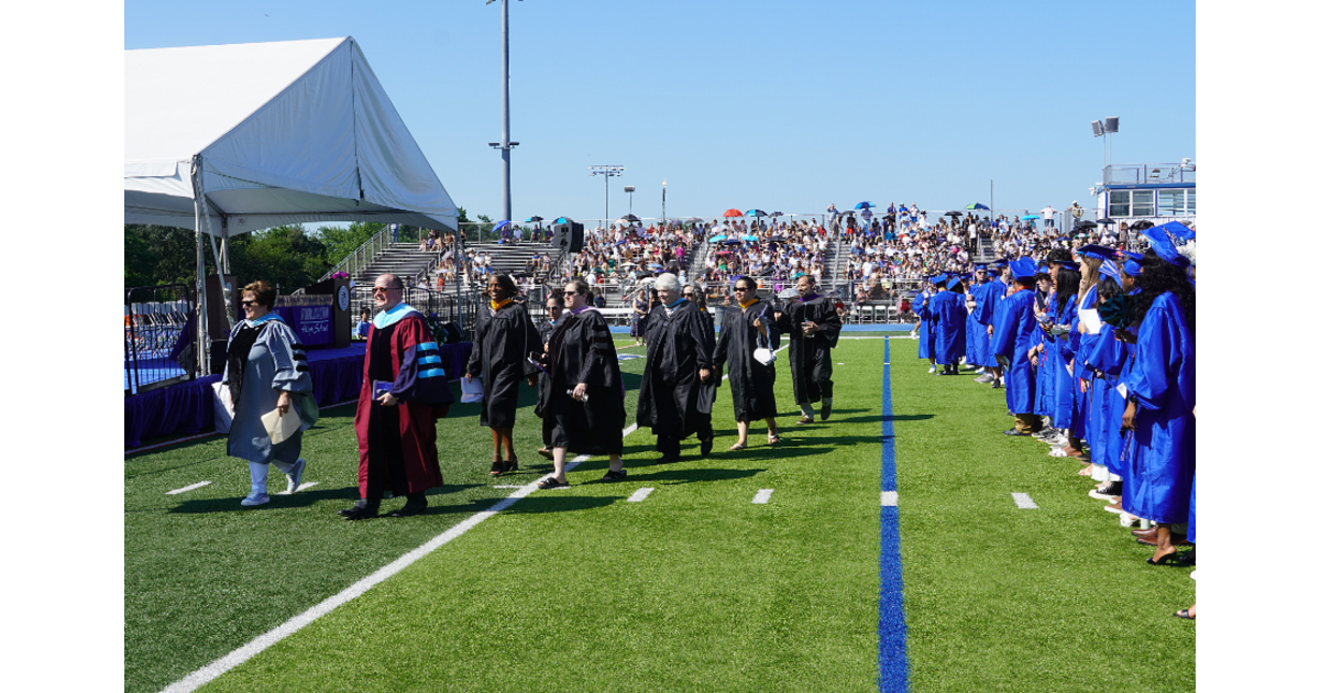 Graduates in caps and gowns walking across a football field at a graduation ceremony, with seated spectators in the background and a large white tent on the left.