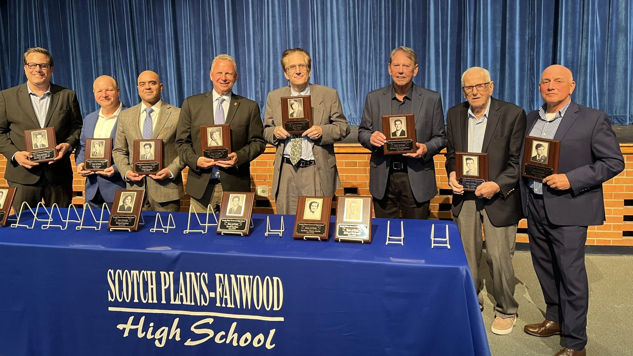 Eight men standing behind a table at an awards ceremony, each holding a framed photo and plaque, with a blue curtain backdrop and a banner that says 'Scotch Plains-Fanwood High School'.