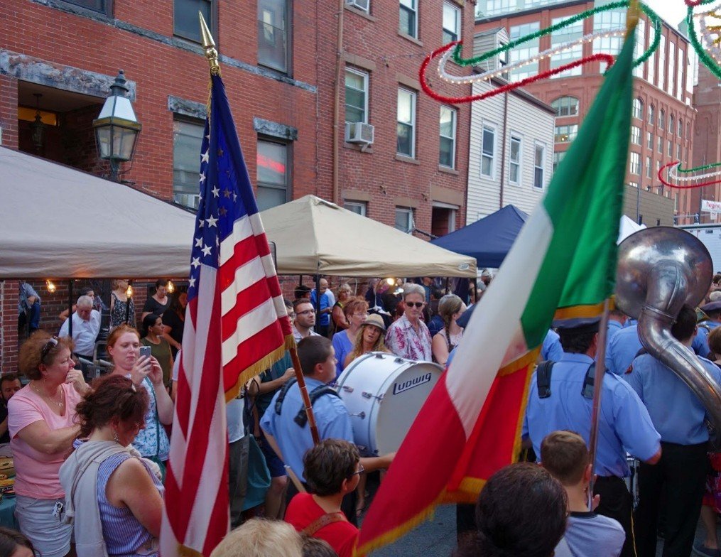 Crowd gathered at an outdoor event with American and Italian flags, a marching band, tents, and string lights, in an urban setting with brick buildings.