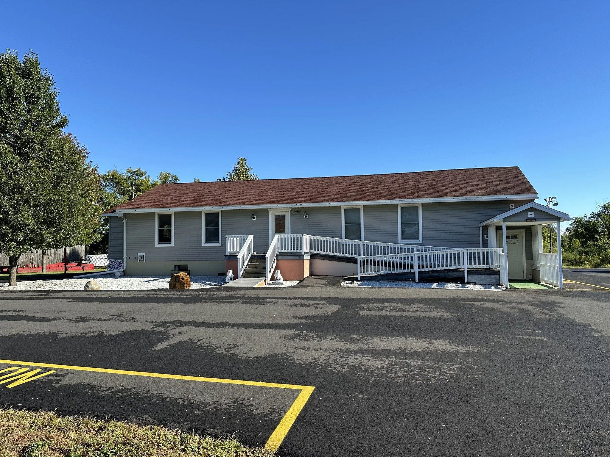 Residential building with a red shingle roof, gray siding, a staircase leading to the front door, and a parking lot with yellow lines and a small patch of grass in the foreground. Trees are on the left side, and the sky is clear and blue.
