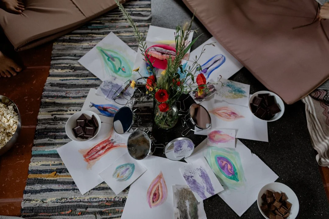 Arrangement of colorful abstract watercolor paintings surrounding a glass vase with red poppies and greenery, with small bowls of dark chocolate pieces and a mirror on a surface covered with a patterned rug and blankets.