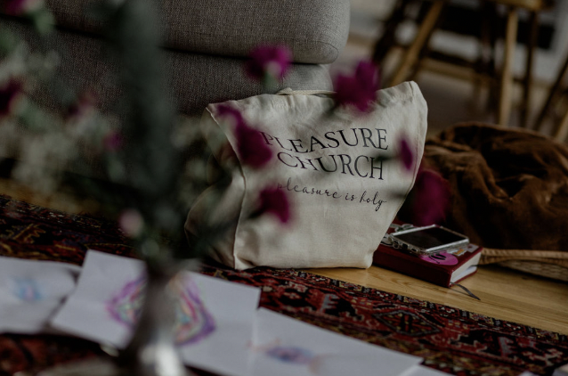 A white pillow with the words 'Pleasure Church' and 'pleasure is holy' written on it, placed on a rug next to a wooden table with a smartphone and some papers.