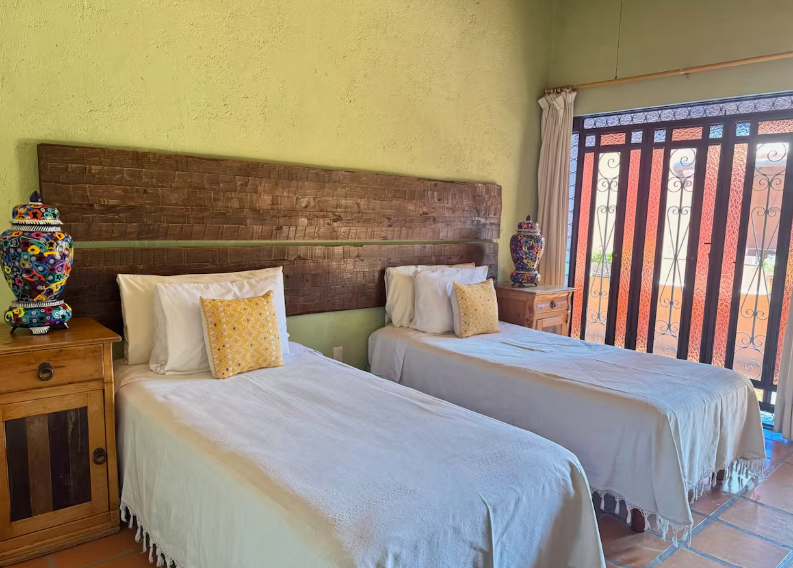 Two twin beds with white linens and decorative yellow pillows, wooden headboards, bedside tables with colorful vases, window with iron grille and curtains, sunlight coming through.