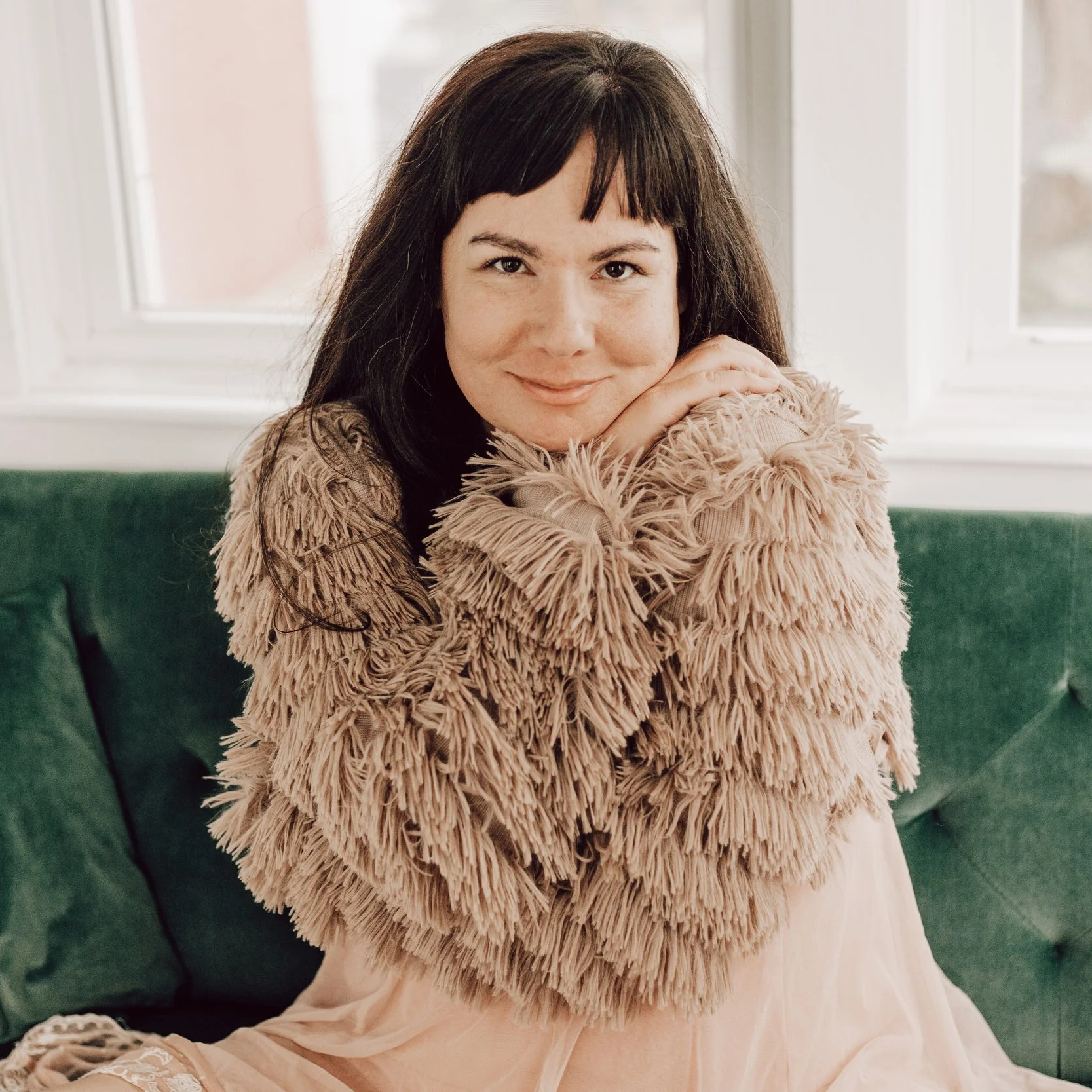 A woman with dark hair, wearing a beige furry coat, sitting on a green sofa near a window.