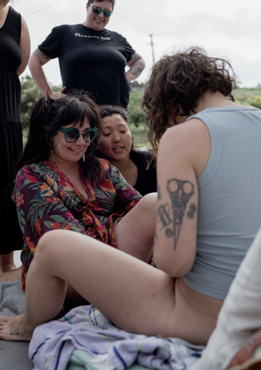 Group of women outdoors, some sitting on a blanket, one wearing colorful clothing and sunglasses, others standing nearby, all smiling and looking at the woman with a tattooed arm showing her knee.