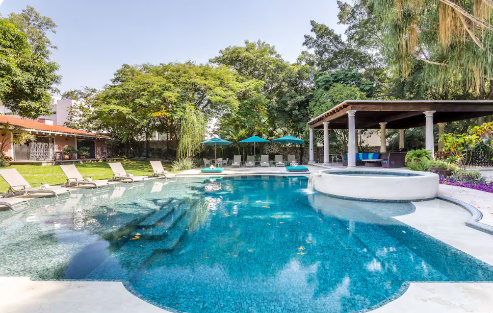 A backyard swimming pool with lounge chairs, umbrellas, and a shaded seating area surrounded by trees and greenery.