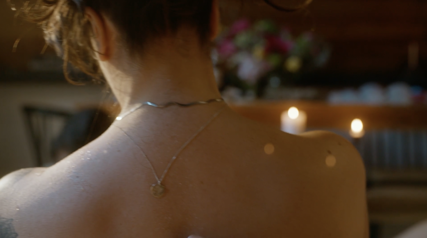 Backside of a woman with wet skin, short hair, wearing a thin chain necklace with a pendant, in a dimly lit room with candles and flowers in the background.