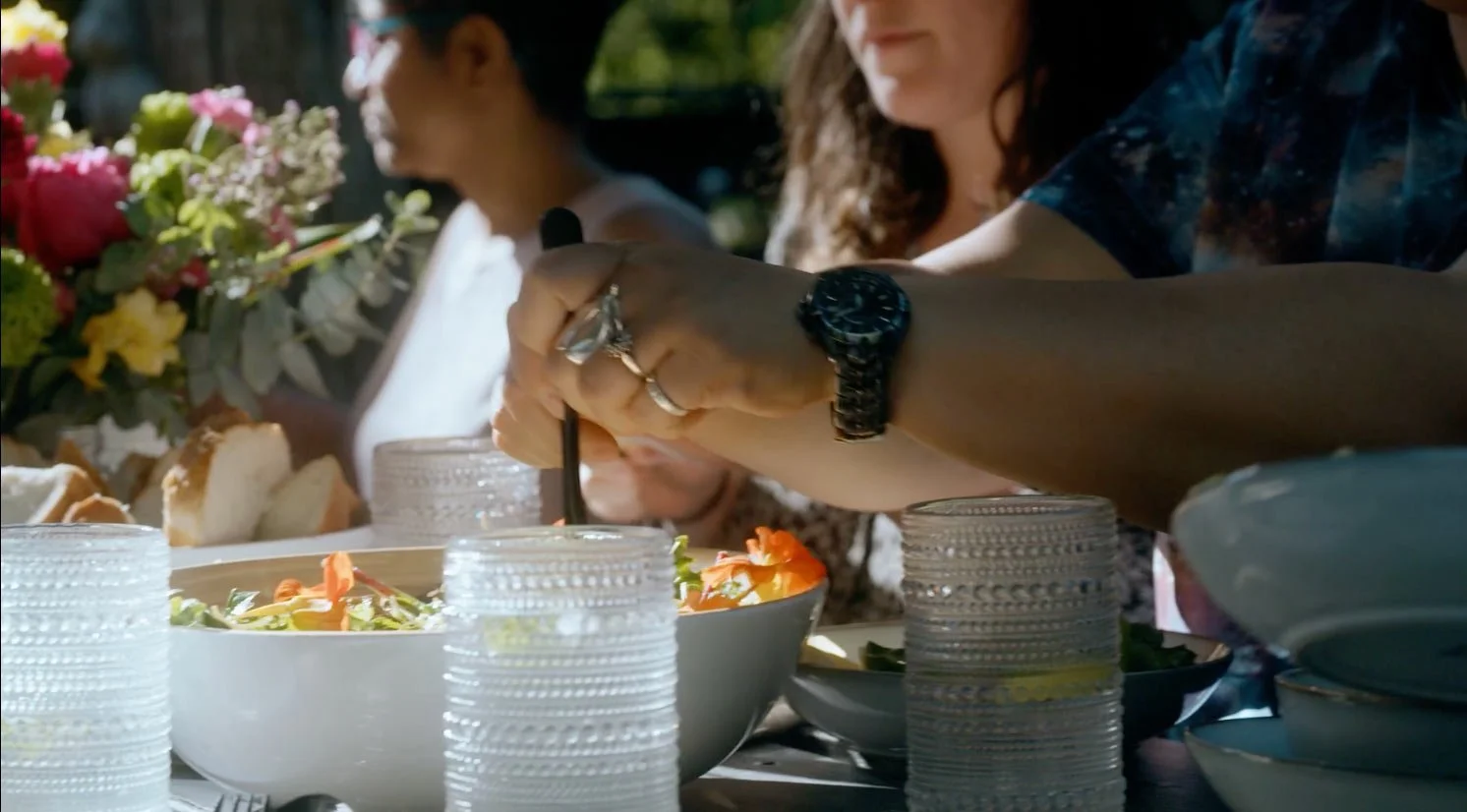 People dining outdoors with salads, bread, and flowers on the table, as one person scoops food from a bowl.