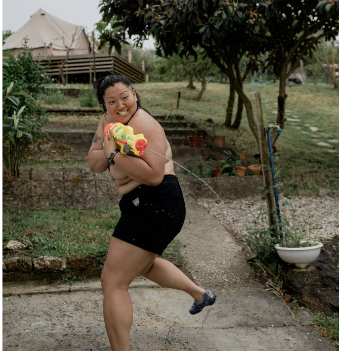 A woman in black shorts and a tattoo on her arm is smiling while playing with a water gun in a garden with trees and potted plants.
