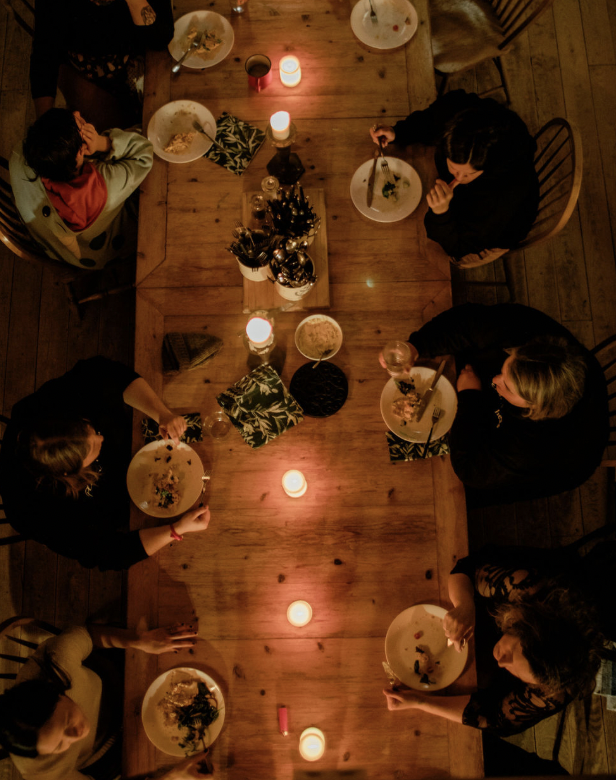 People sitting around a wooden dining table enjoying a meal, decorated with candles and a small Christmas tree in the center, in a warmly lit room.