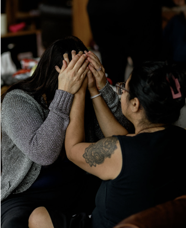 Two women sitting close together, one woman with dark hair covering her face with her hands, being comforted or consoled by the other woman with a tattooed arm, glasses, and a hair clip, in an indoor setting.