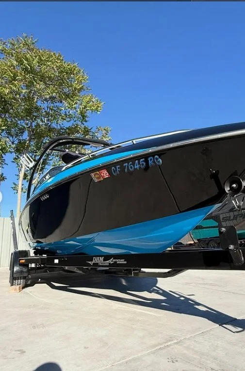 A black and blue speedboat on a trailer in an outdoor lot at sergio's boat spa shop, with a clear blue sky and a tree in the background.