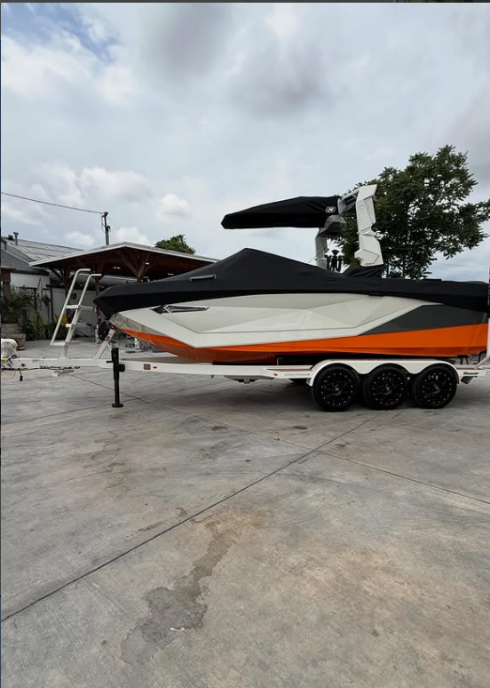 A speedboat on a trailer parked on a concrete driveway in a residential area with houses, trees, and a cloudy sky in the background.