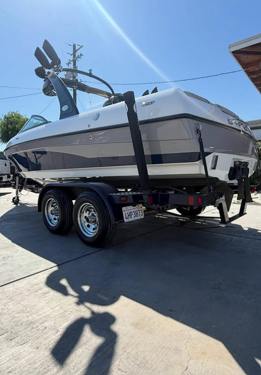 A white and navy speedboat on a trailer parked on a concrete driveway, with a shadow cast on the ground and a building partially visible on the right.