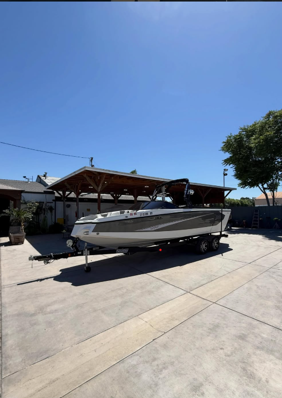 A white and gray speedboat on a trailer parked on a concrete driveway with a wooden carport, house, and tree in the background under a clear blue sky.