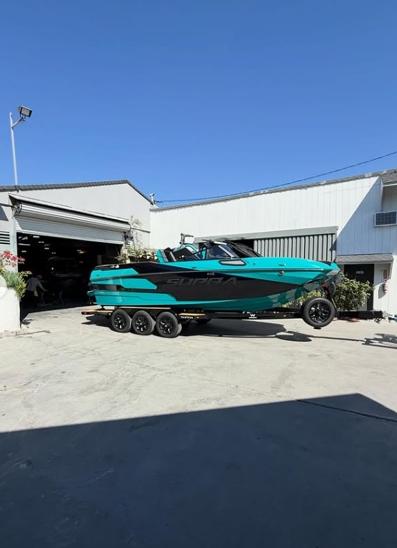 A teal and black Supra speedboat on a triple-axle trailer outside a white industrial building under a clear blue sky.