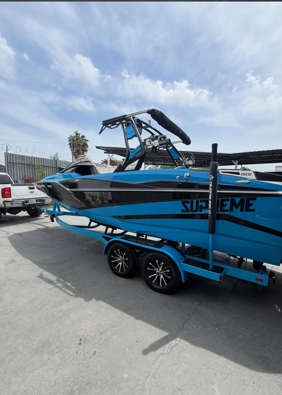A blue and black SUPREME branded wakeboard boat on a trailer in a parking lot under a partly cloudy sky.