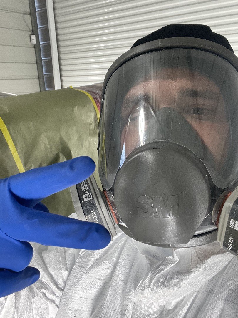 Person wearing a full face respirator mask and blue gloves holding an air filter canister in front of a metal garage door.