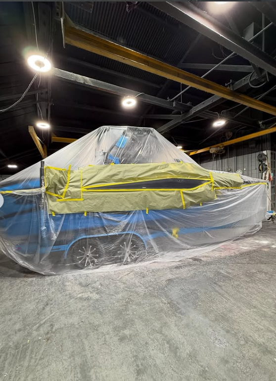 A Boat covered with a large plastic and painter's tape, inside a workshop or garage with overhead lighting and industrial infrastructure.