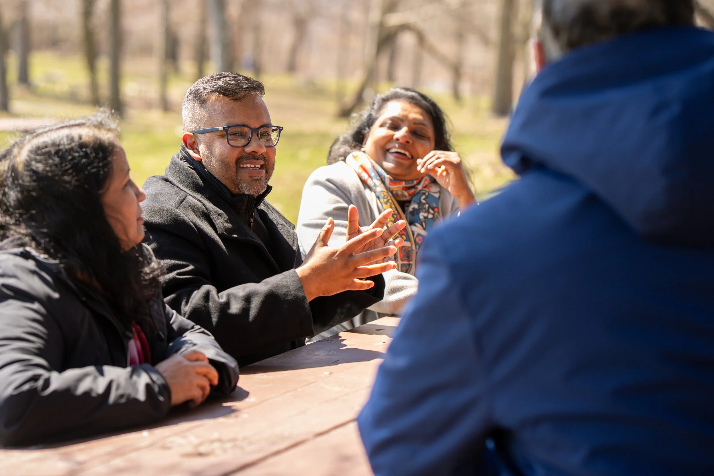 Group of people sitting at an outdoor table, engaged in conversation, in a park with trees in the background on a sunny day.