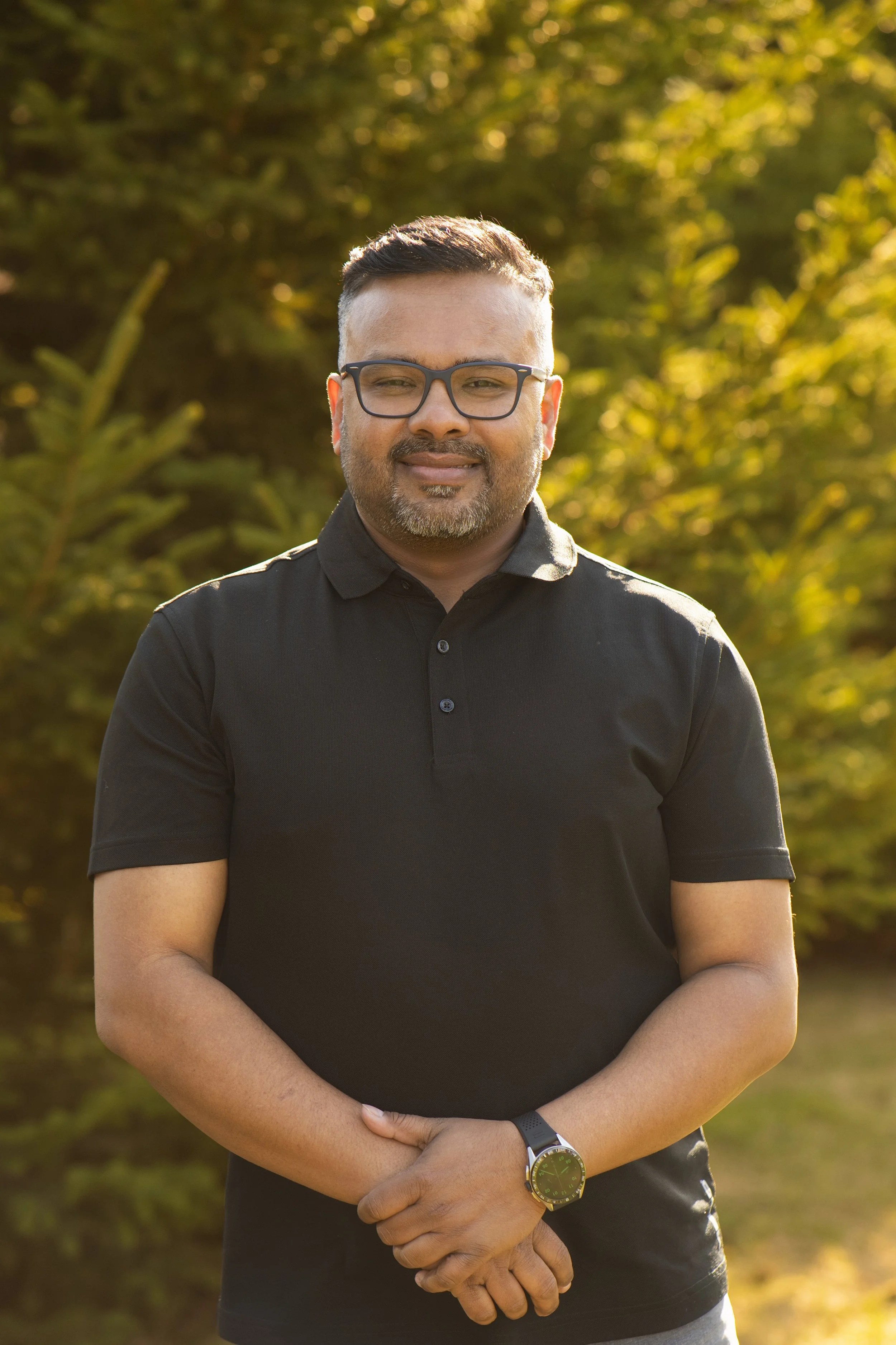 A man with glasses and a beard wearing a black polo shirt standing outdoors in front of green trees during the daytime.