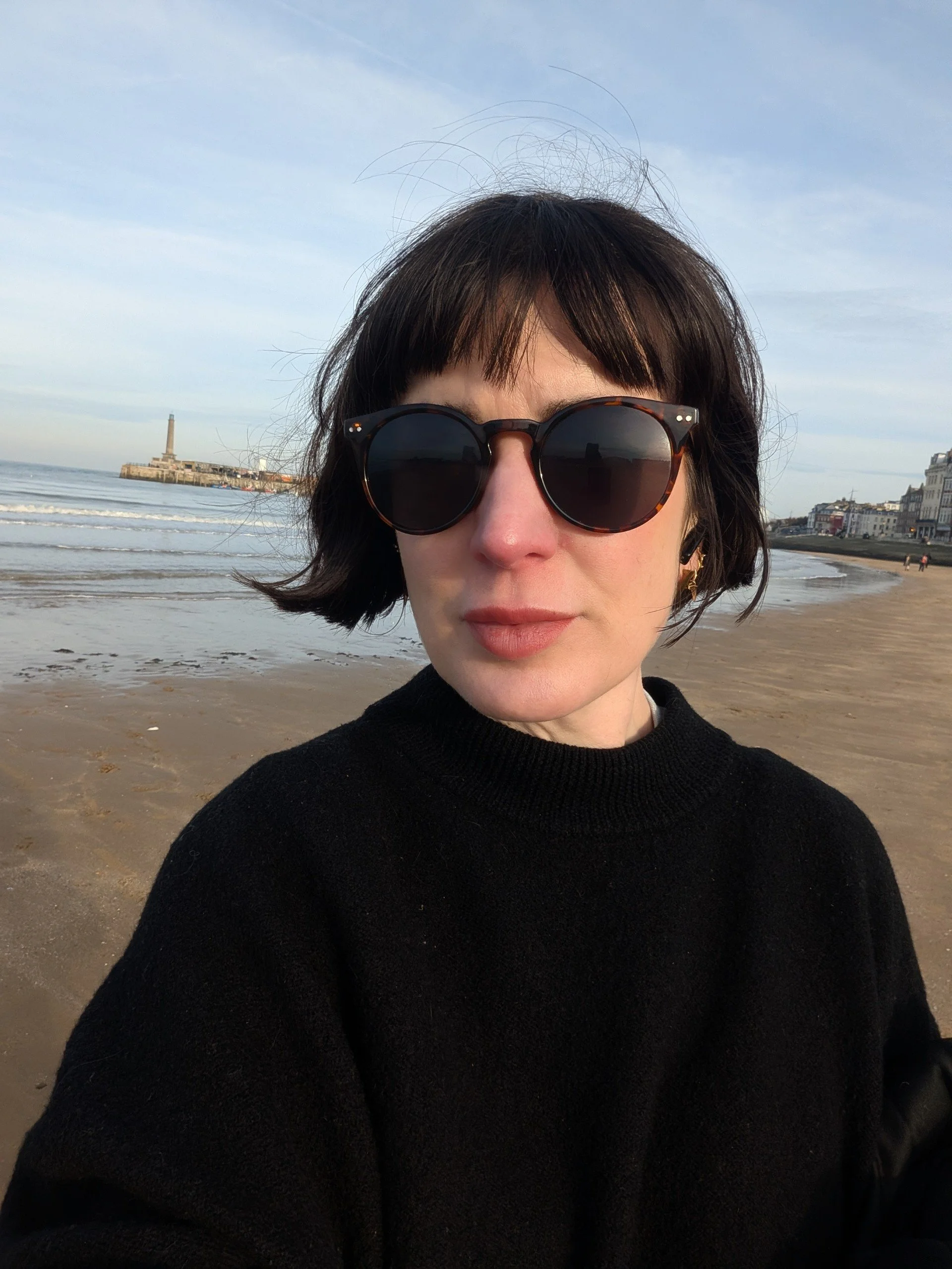 Close-up of a woman with short dark hair and sunglasses taking a selfie on a beach with a lighthouse and buildings in the background.