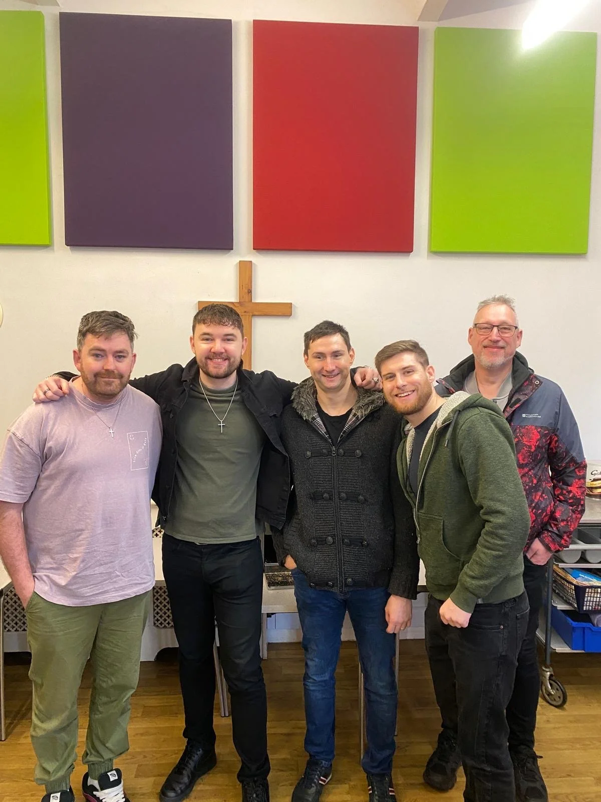 Group of five men standing together indoors, some with arms around each other, in front of a cross and colorful wall art.