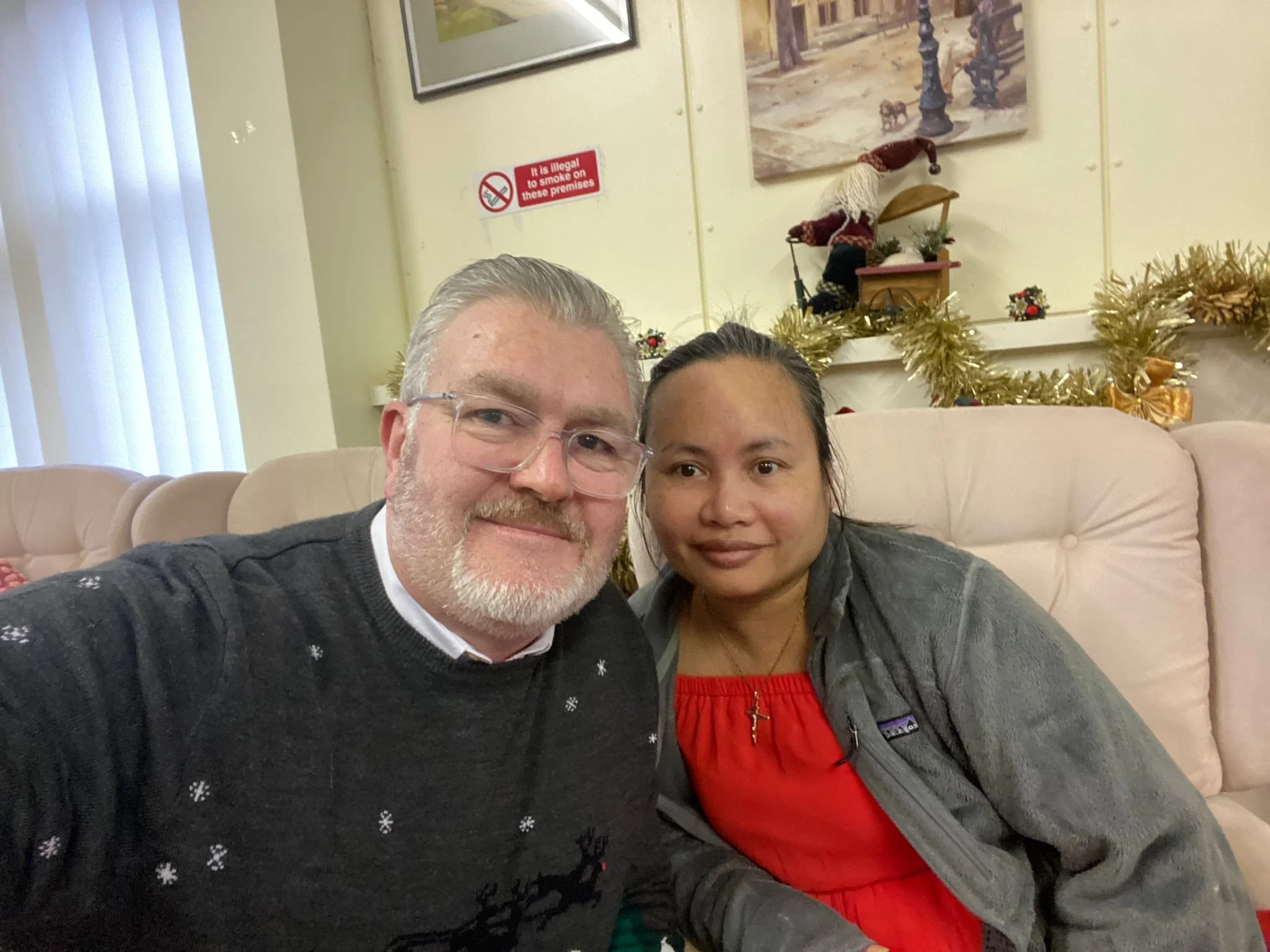 A selfie of a man with glasses and a beard and a woman with dark hair in a ponytail, sitting on a beige sofa in a decorated living room with Christmas decorations. There is a red sign on the wall indicating it is illegal to smoke on these premises, and a painting and a tan-colored curtain are also visible in the background.
