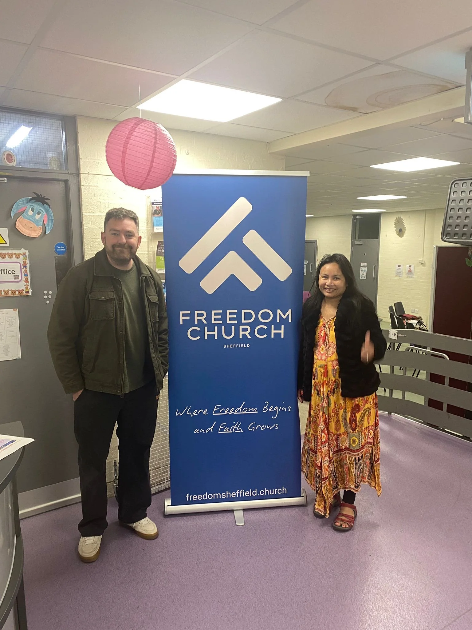 Two people standing next to a blue banner with the words 'Freedom Church Sheffield' and the slogan 'Where Freedom Begins and Faith Grows.' They are in an indoor space with a pink paper lantern hanging from the ceiling, a door, and various decorations in the background.