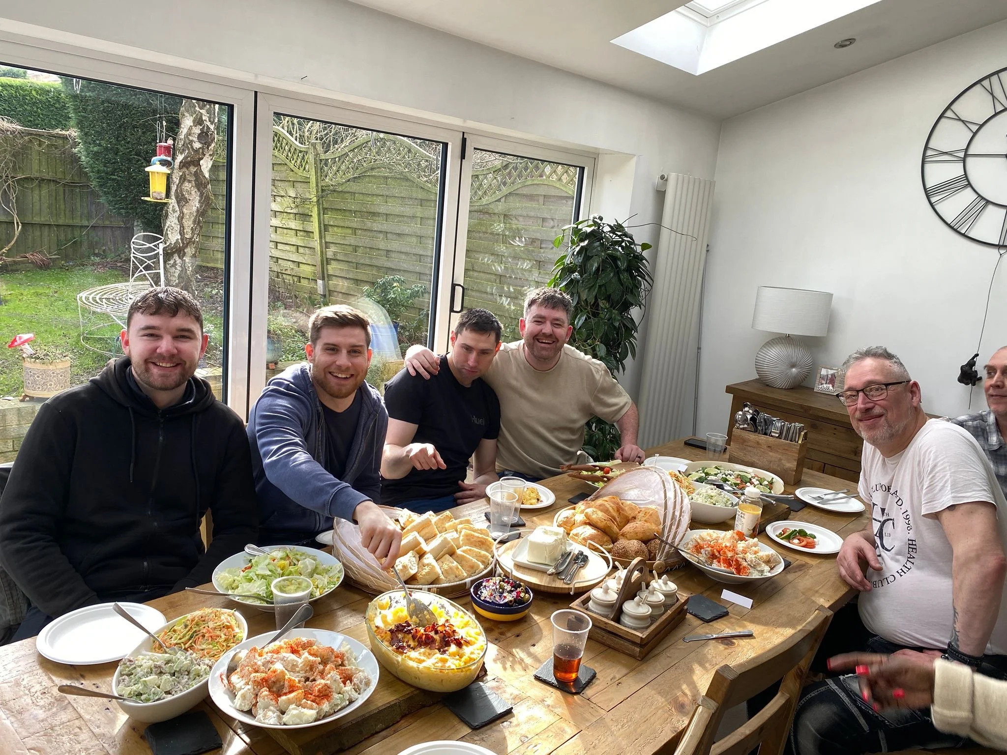 Group of men sitting around a wooden table set with various salads, bread, and desserts during a meal in a bright room with large windows and sliding glass doors leading to a backyard.