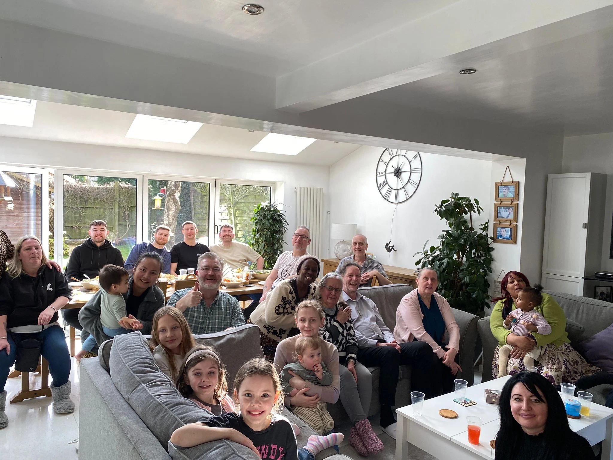 A group of people gathering in a bright, modern living room with large windows, a wall clock, and indoor plants, celebrating a social event or family gathering.