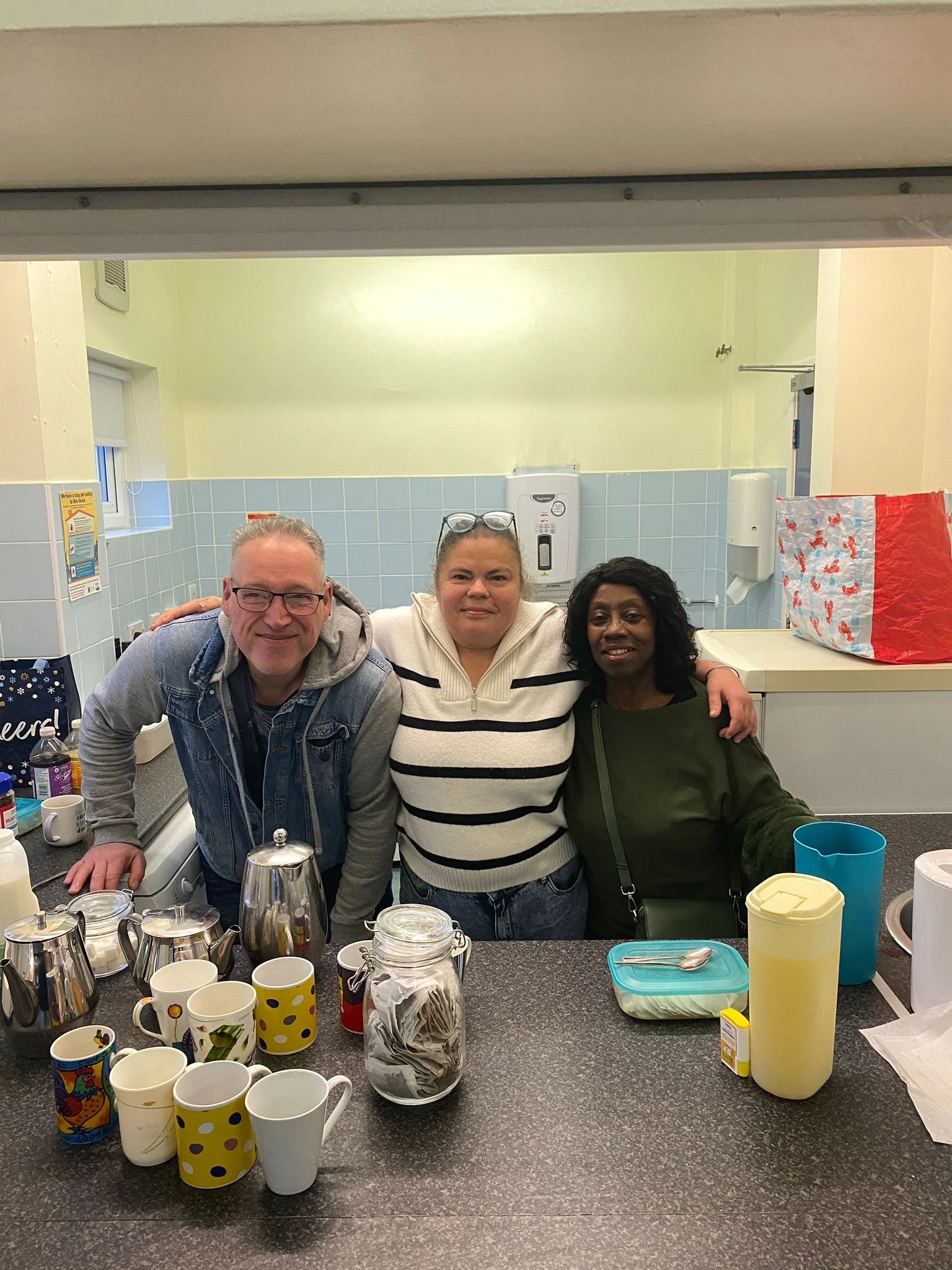 Three people smiling and posing for a photo in a kitchen or break room, with coffee mugs, bottles, and containers on the counter in front of them.