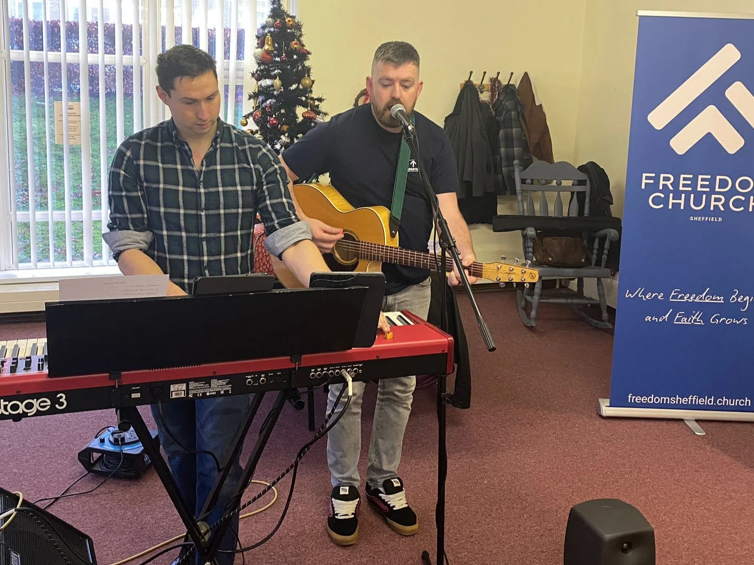 Two men performing music indoors, one playing a keyboard and the other singing and playing an acoustic guitar, near a decorated Christmas tree and a banner for Freedom Church Sheffield.