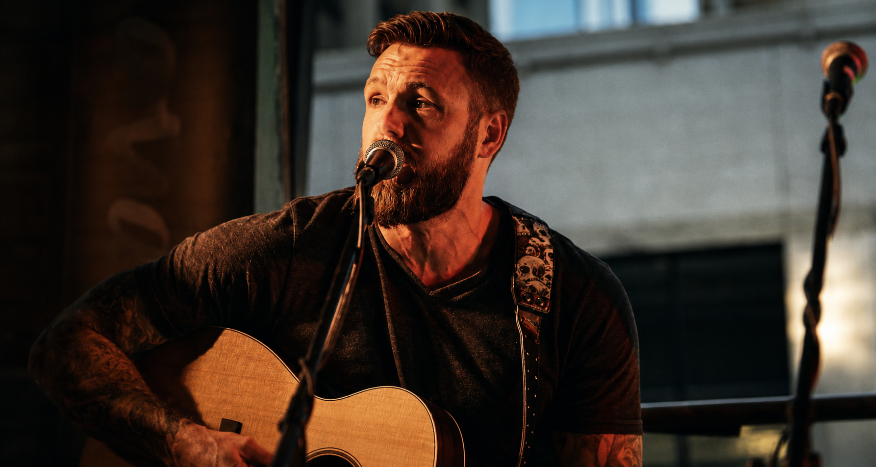 A man with a beard and tattoos playing an acoustic guitar and singing into a microphone during a performance.
