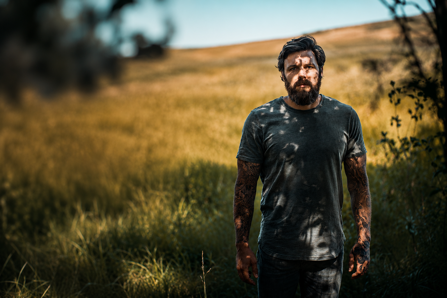 A man with dark hair and a beard, wearing a black t-shirt with a camouflage pattern, standing in a grassy outdoor setting with trees and a hill in the background.