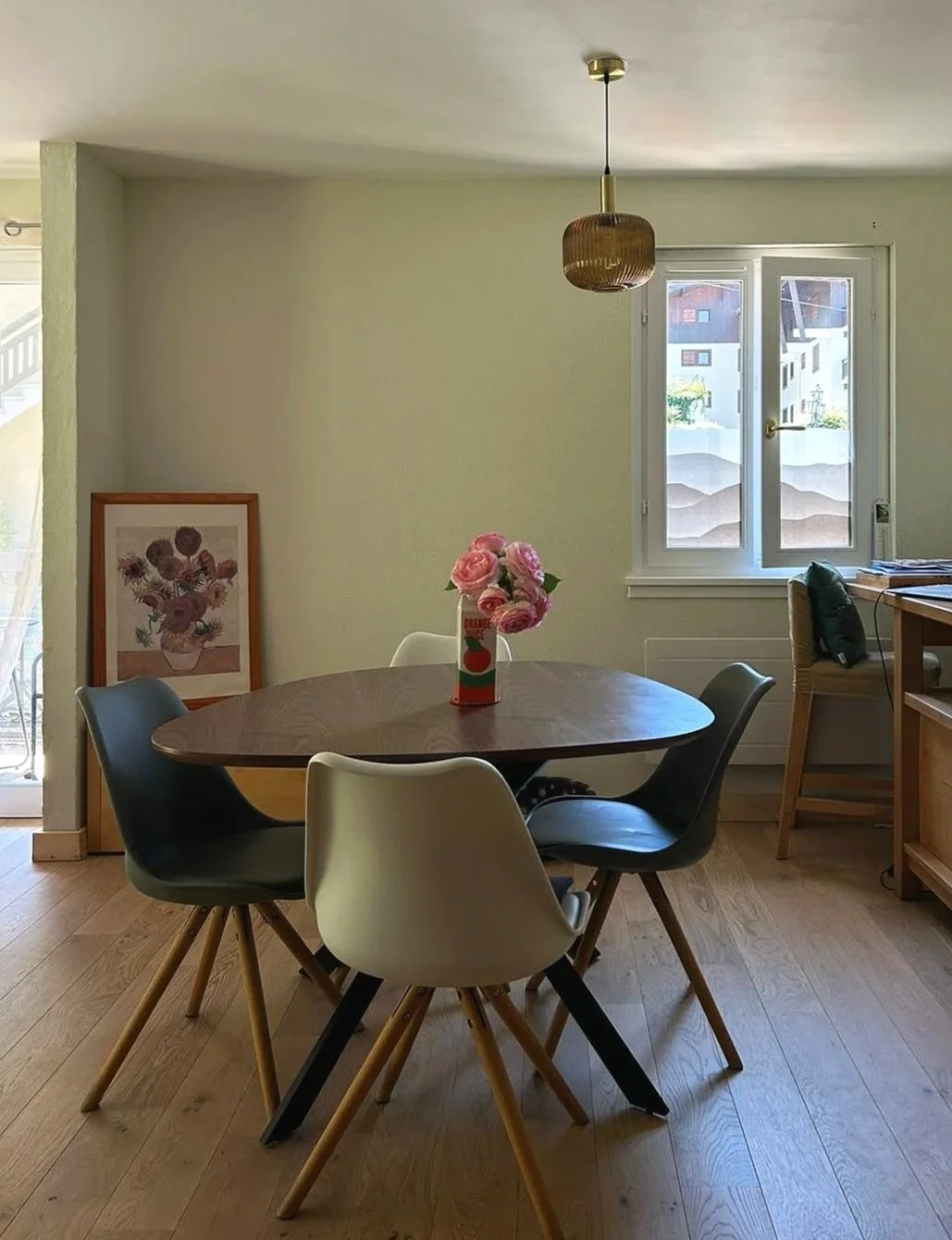 Salle à manger avec un mur blanc, un tableau de fleurs en cadre, une table ronde en bois avec un vase de fleurs roses, des chaises modernes autour, une fenêtre laissant entrer la lumière, un plafond avec une suspension.