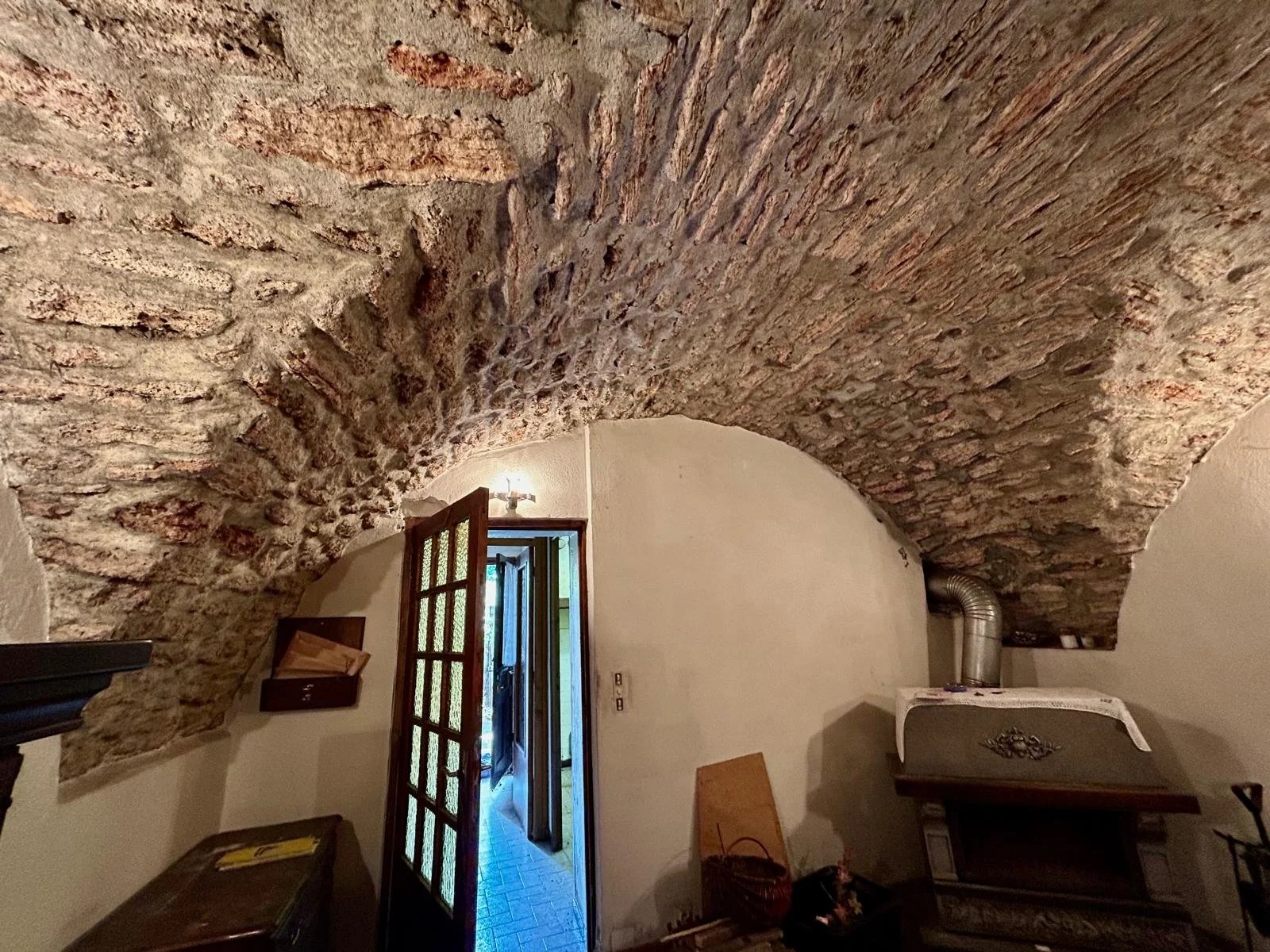 Intérieur d'une pièce avec un plafond en voûte en briques apparentes, une porte en bois ouverte laissant entrer de la lumière, et quelques meubles anciens.