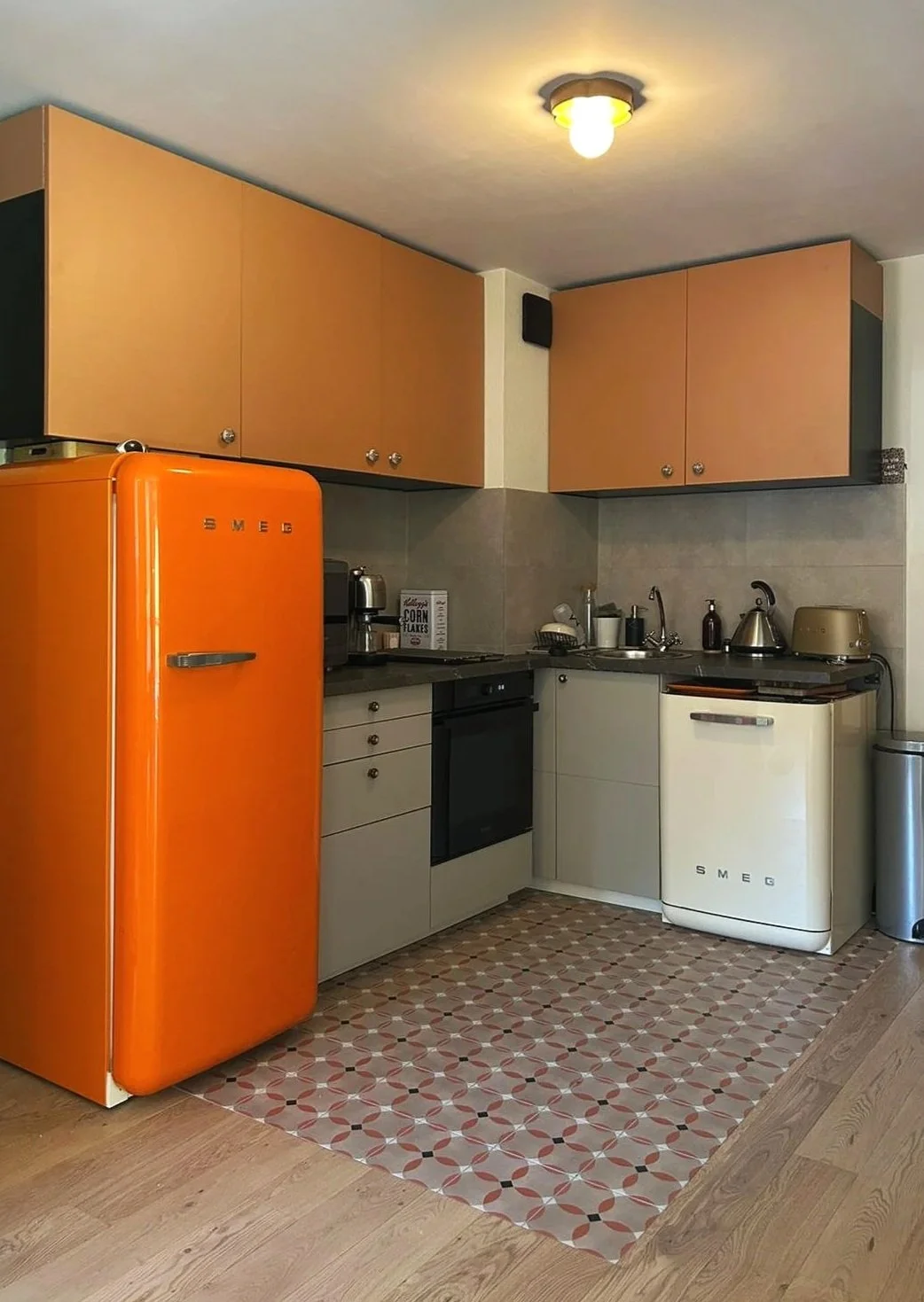 Cuisine avec un réfrigérateur orange, un four noir, des armoires en bois clair, une évier, une machine à laver Smeg blanche, une bouilloire en métal, un grille-pain doré, et un tatami de motifs géométriques en rouge et beige.