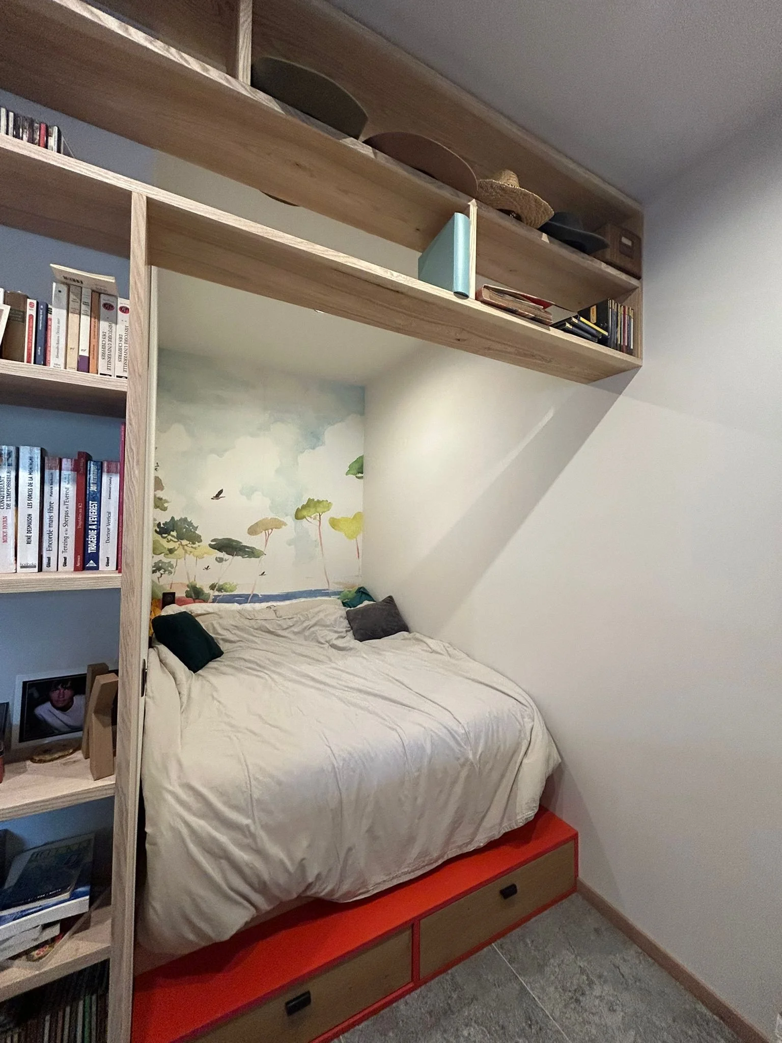 Une chambre avec un lit avec draps blancs près d'une étagère en bois contenant des livres et des objets, avec un mur en arrière-plan décoré d'une peinture ou d'une fresque représentant un paysage avec des arbres et un ciel.