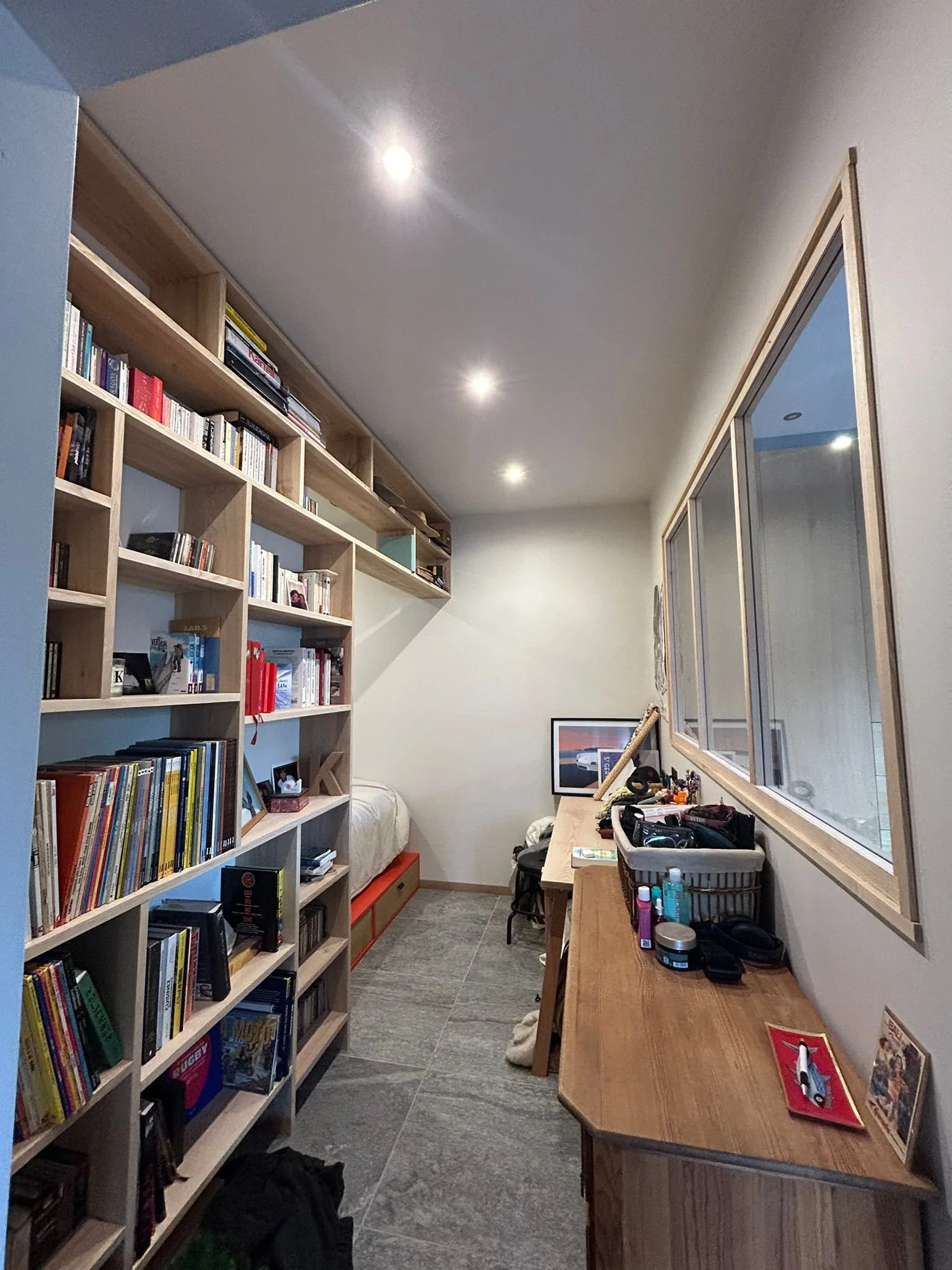 Une petite pièce de maison ou d'appartement avec une grande étagère en bois remplie de livres, un bureau avec divers objets et une fenêtre en longueur côté droit.