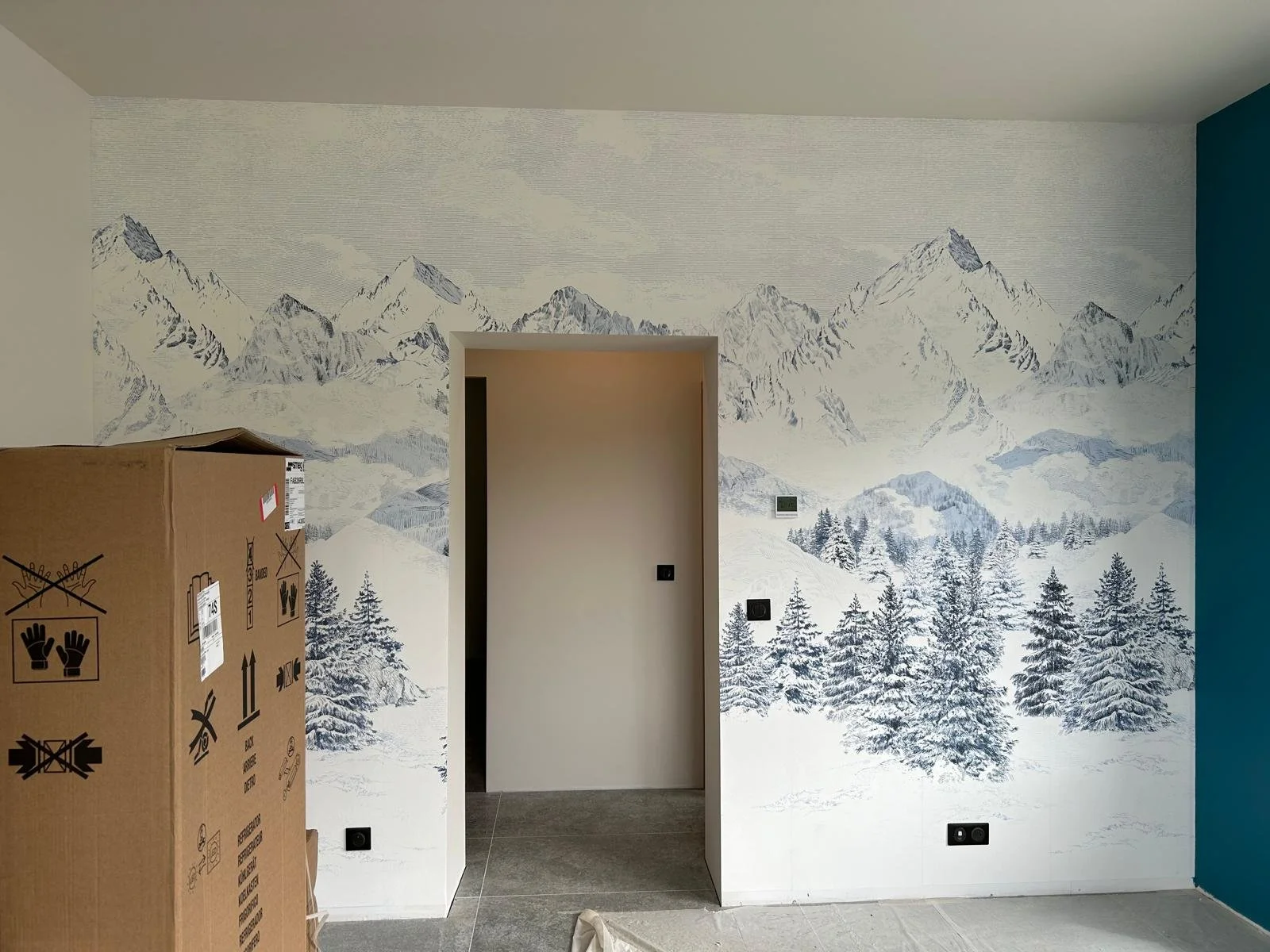 Mur en papier peint peint représentant une scène montagneuse enneigée avec des arbres et un ciel nuageux, dans une pièce en cours de décoration.