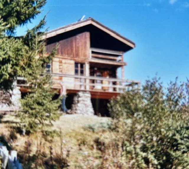 Maison en bois située sur une colline, entourée d'arbres, sous un ciel bleu.