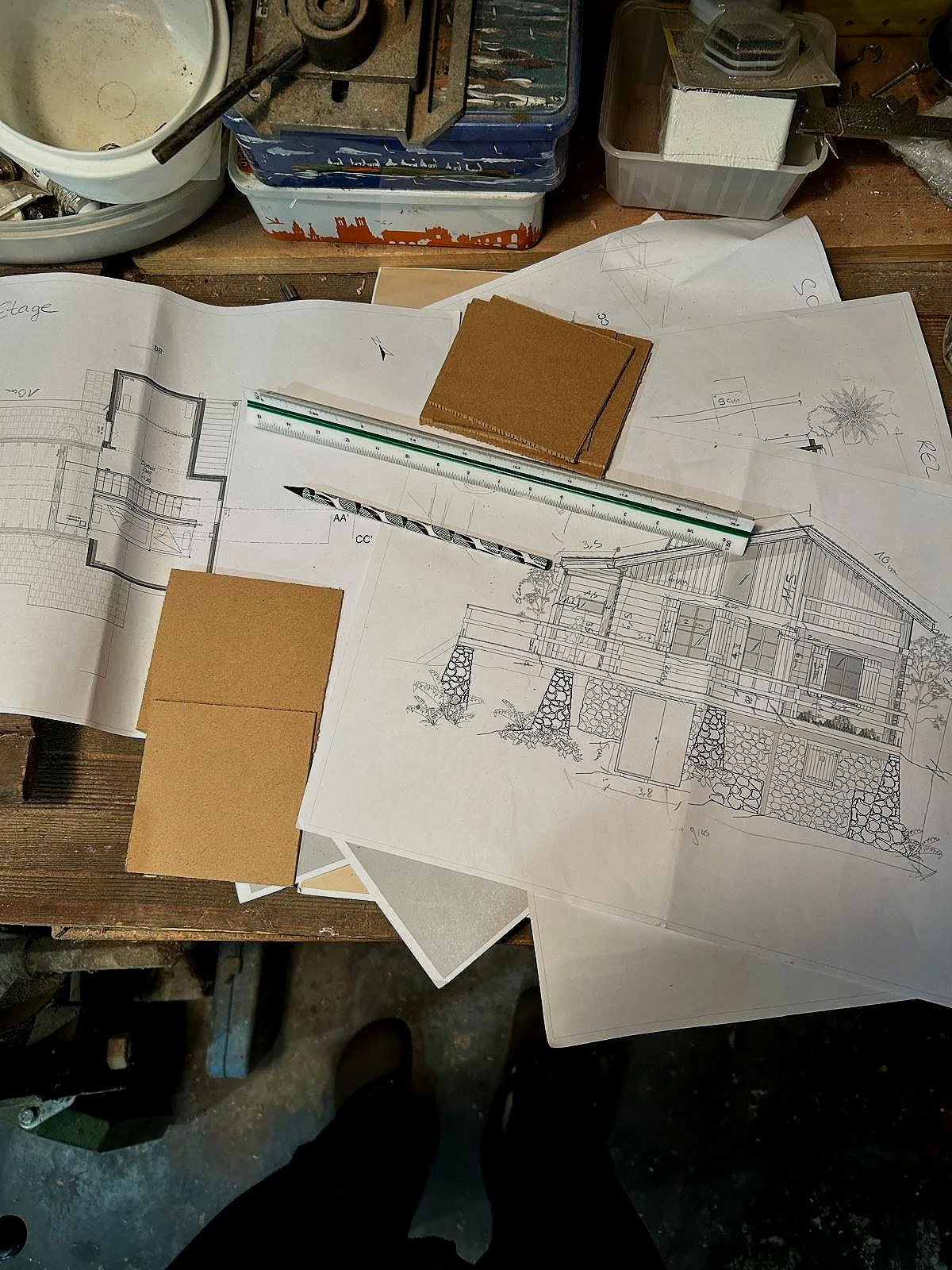 Plans d'architecture et échantillons de matériaux posés sur une table de travail, avec des outils et des boîtes de rangement autour.