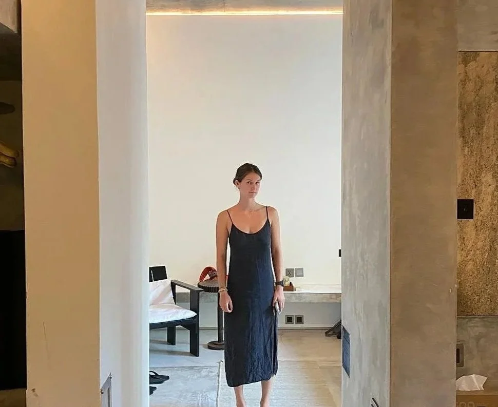 Une femme en robe noire debout dans une pièce avec un décor moderne, dans une optique ouverte entre deux murs.