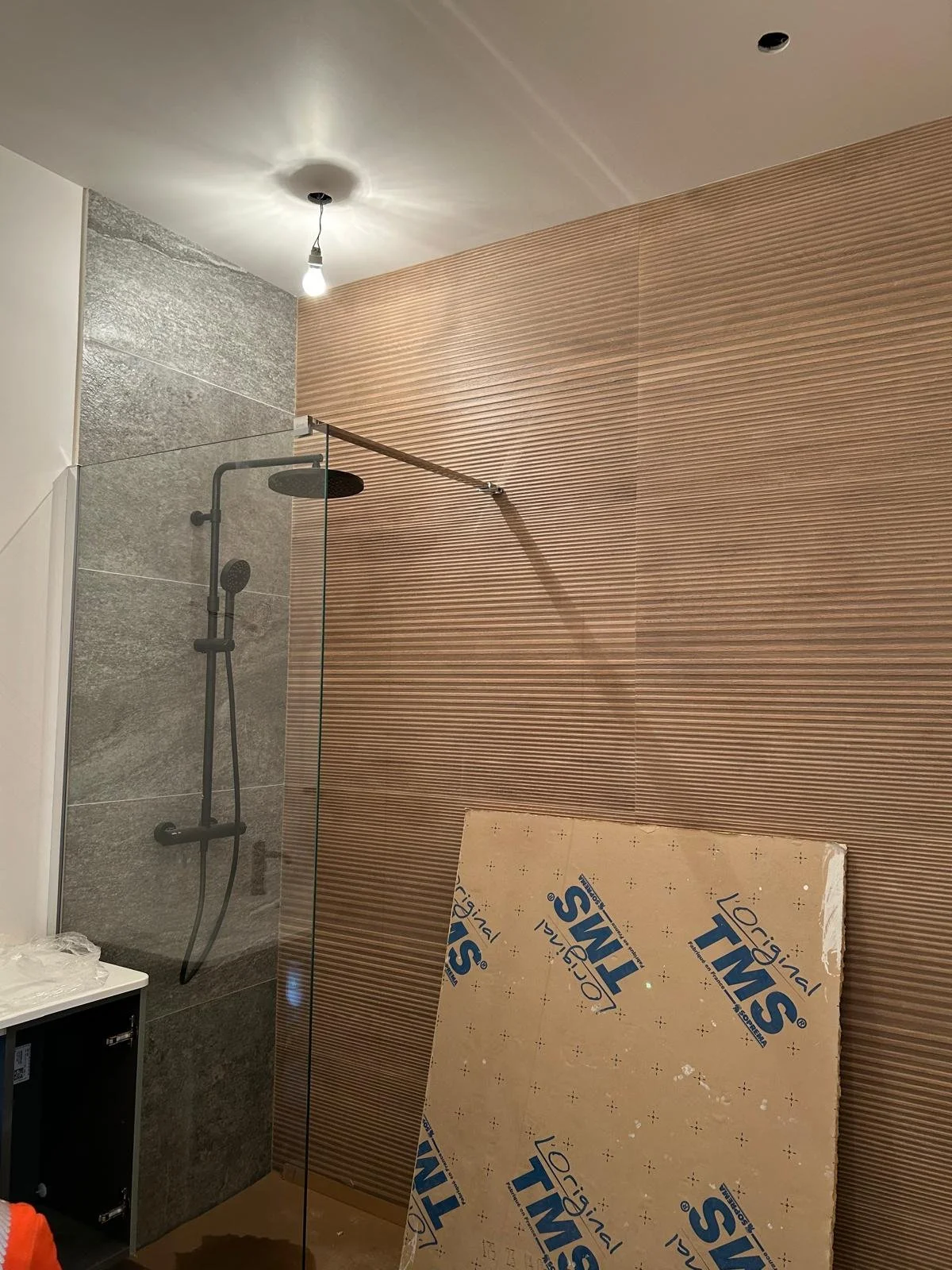 Une cabine de douche en cours d'installation avec un mur en carreaux de pierre gris et un mur en lamelles de bois, une plaque de matériau insonorisant posée au sol, un plafonnier électrique allumé, une colonne de douche noire et une paroi de verre partielle.