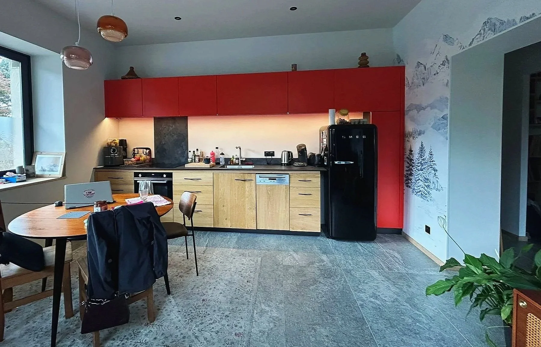 Cuisine moderne avec plaques de cuisson, réfrigérateur noir de marque SMEG, plan de travail avec divers appareils électroménagers, table en bois avec des chaises, décoration murale avec une scène de montagne, fenêtre laissant entrer la lumière, plante verte dans un pot en osier.