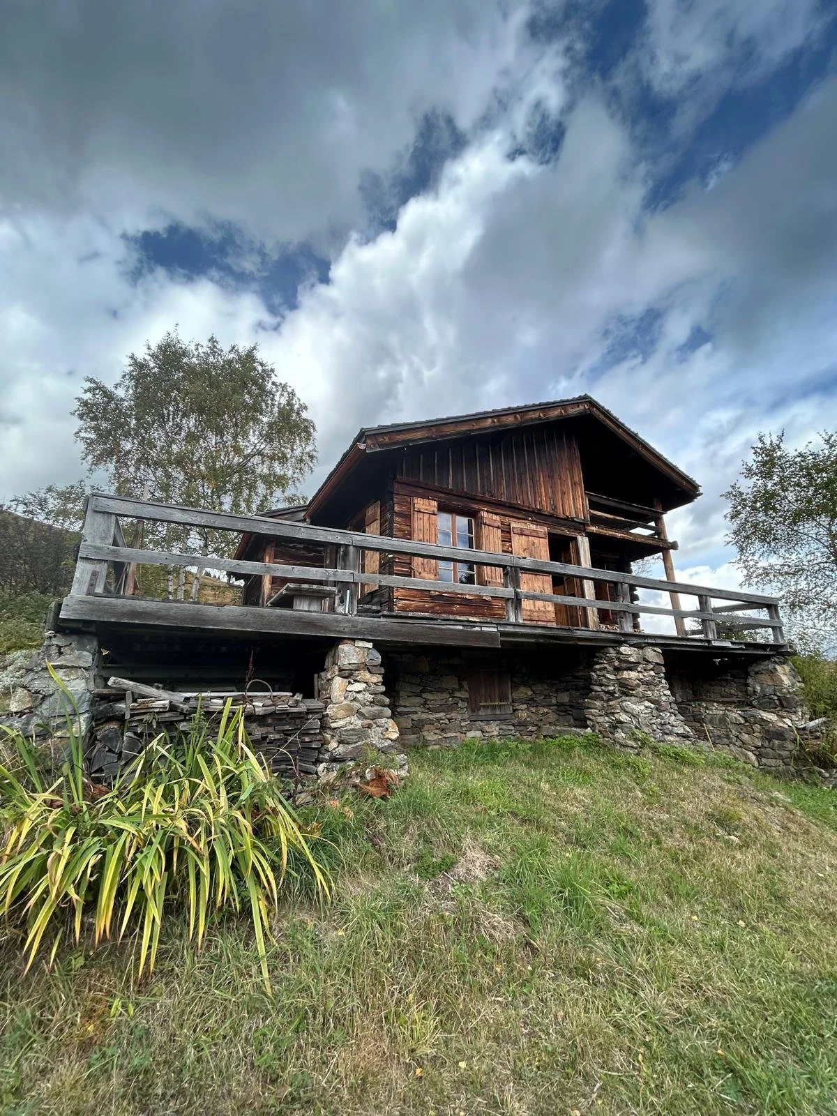 Une maison en bois située sur une fondation en pierres, avec un balcon en bois, entourée de verdure et de deux grands arbres, sous un ciel partiellement nuageux.